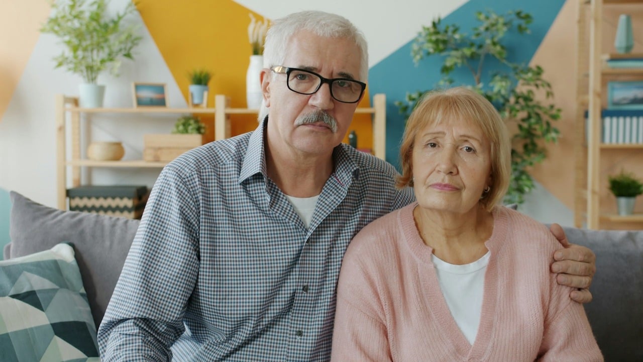 An older couple sitting on a couch with the man's arm around the woman's shoulder.