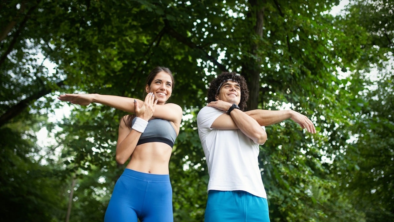 Man and woman stretching their arms outdoors with a background of trees.