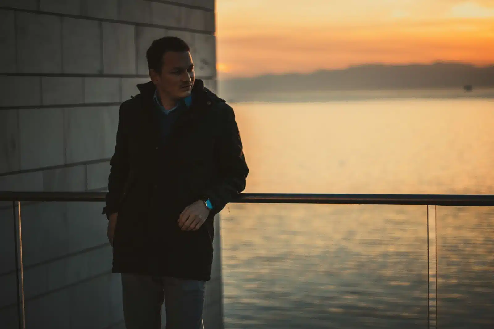 A man standing alone leaning against a railing at sunset