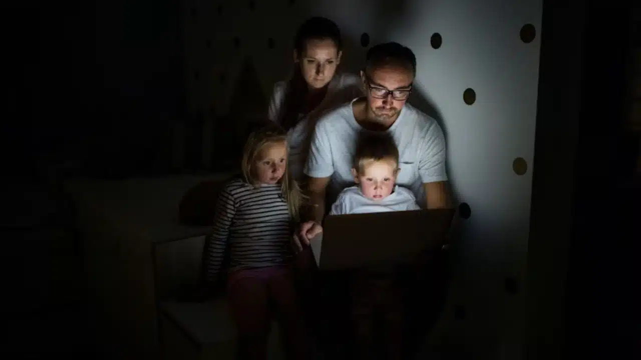 Man working on laptop late at night while family is visible nearby