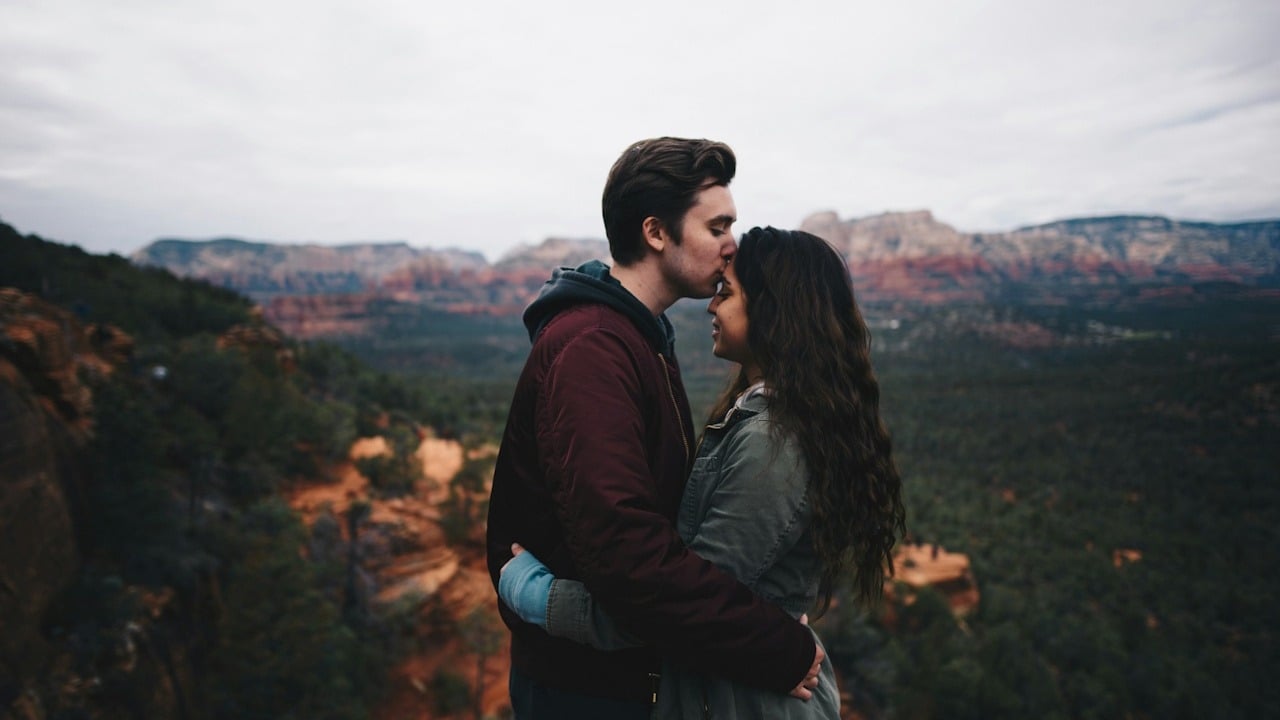 A couple embracing, with the man kissing the woman's forehead, against a backdrop of sweeping red rock mountains under a cloudy sky.