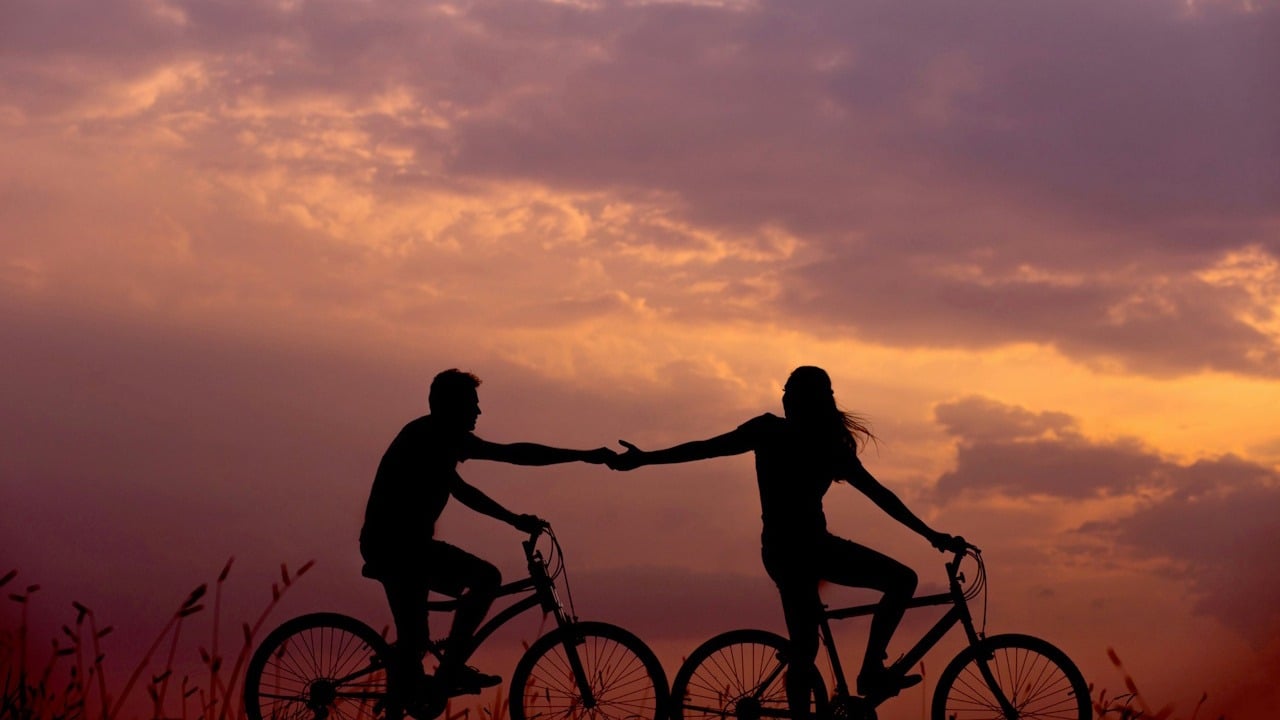 A silhouette of a couple riding bicycles against a vivid orange and purple sunset sky, reaching out to hold hands.