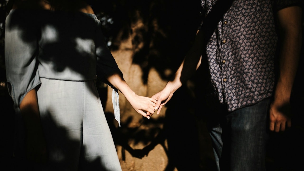 Close-up of a couple holding hands in dim lighting with dark shadows cast on a textured wall.