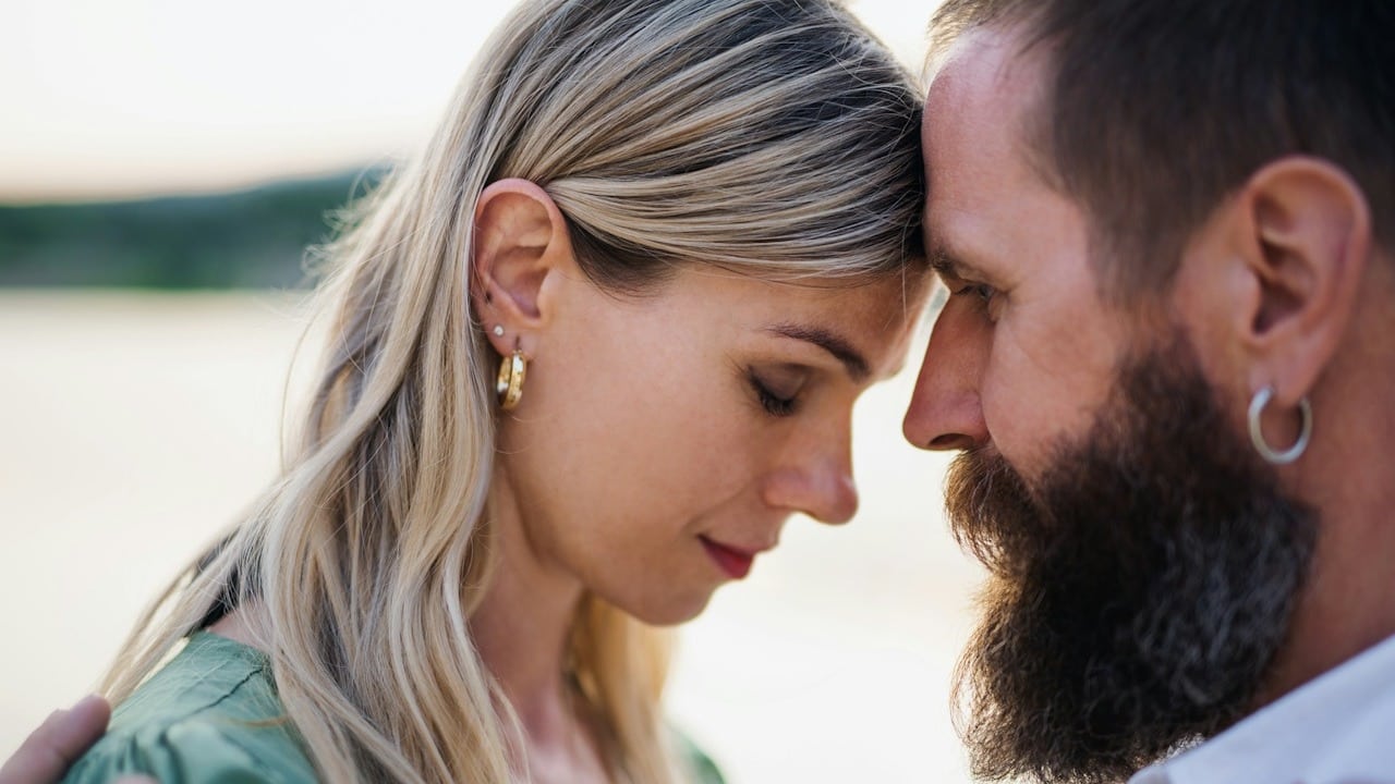 A close-up of a bearded man and a blonde woman with their foreheads touching in an intimate moment outdoors.