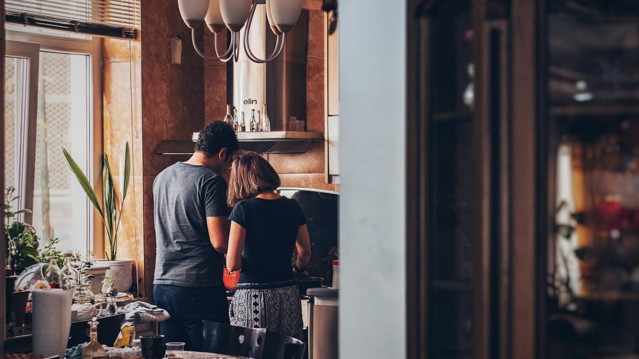 A couple stands close together in a kitchen with their backs to the camera as they cook together.