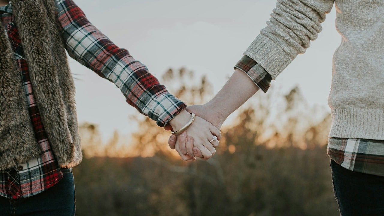 Close-up of a couple holding hands outdoors, with the sunset or golden hour light visible in the blurred background.