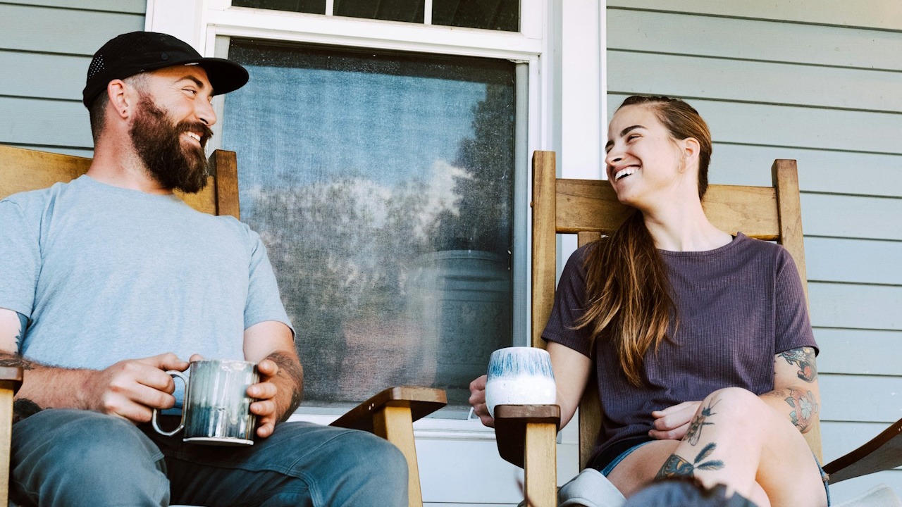 A couple smiling while having coffee on a porch.