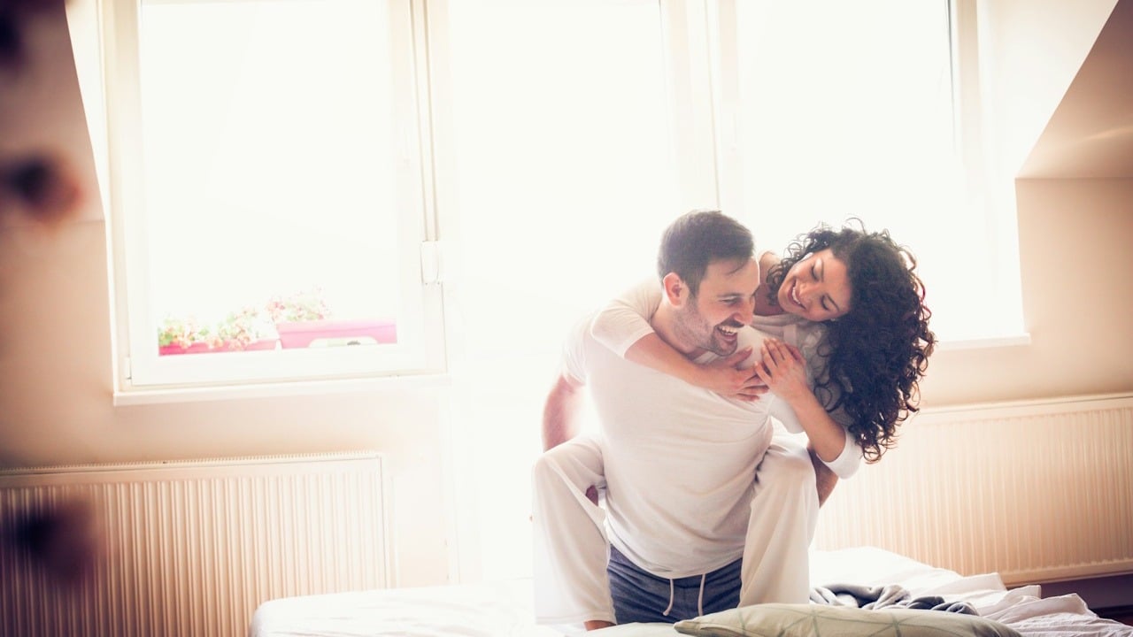 A man in white pajamas is giving a piggyback ride to a woman with curly hair on a bright bed in front of a large window.