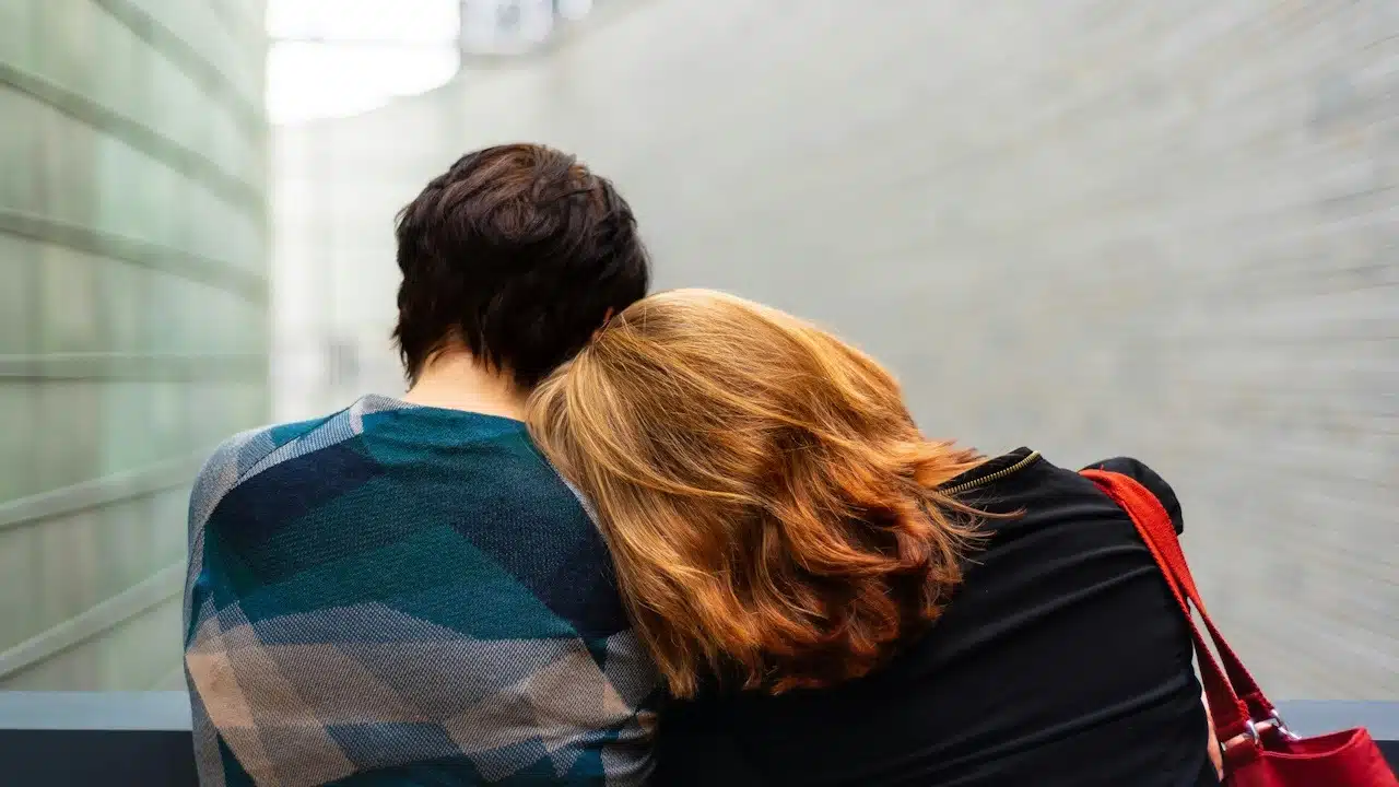 Back view of two people, one with their head resting on the other's shoulder.