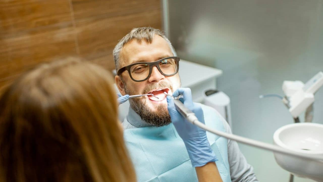 Man in glasses with a beard smiles as a dentist in blue gloves examines his teeth.