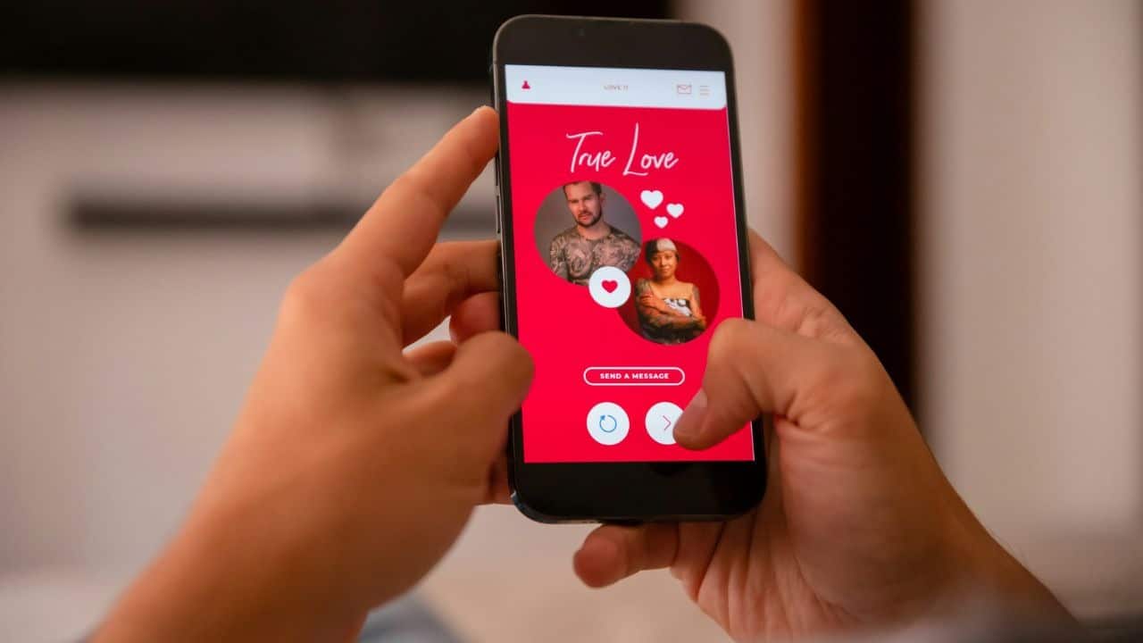Two hands hold a smartphone displaying a red dating app interface with two user profiles.