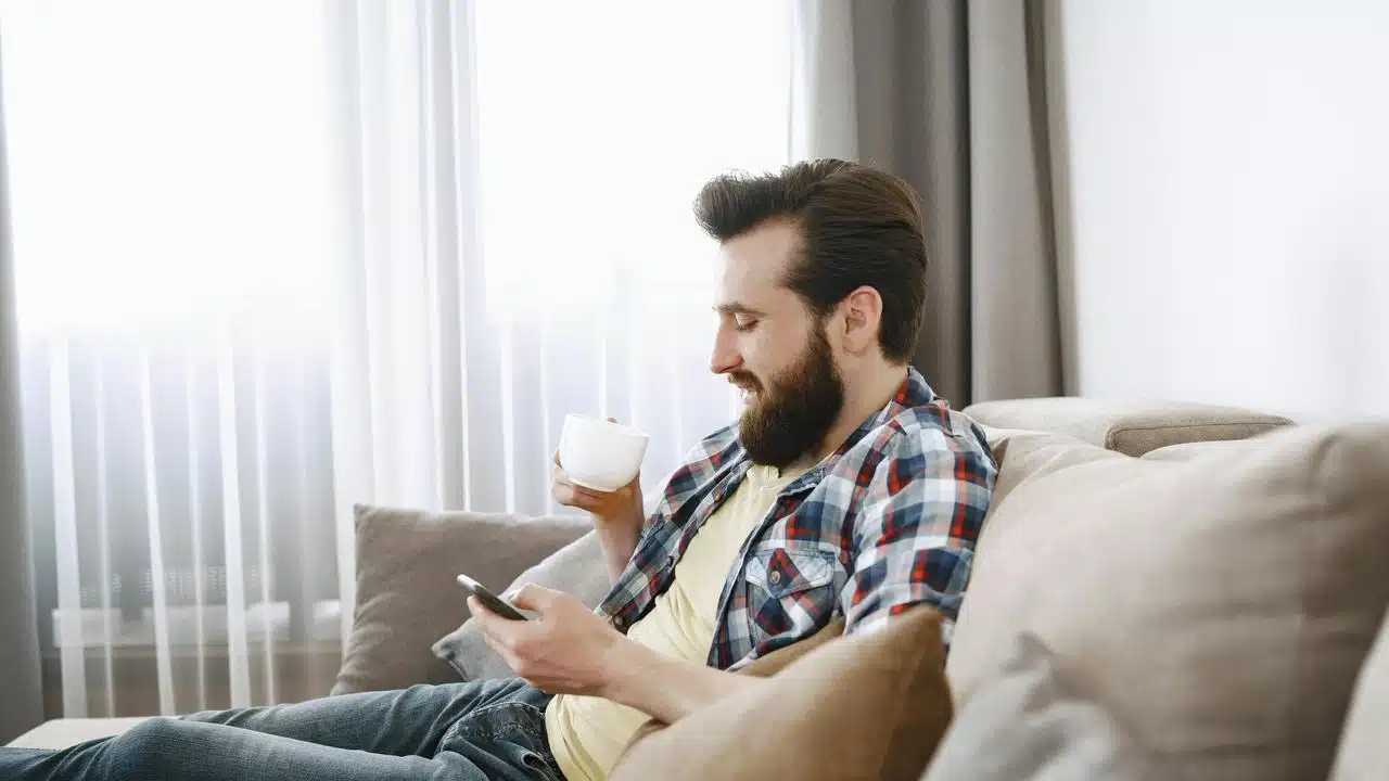 A bearded man sitting on a couch using his phone and holding a cup.