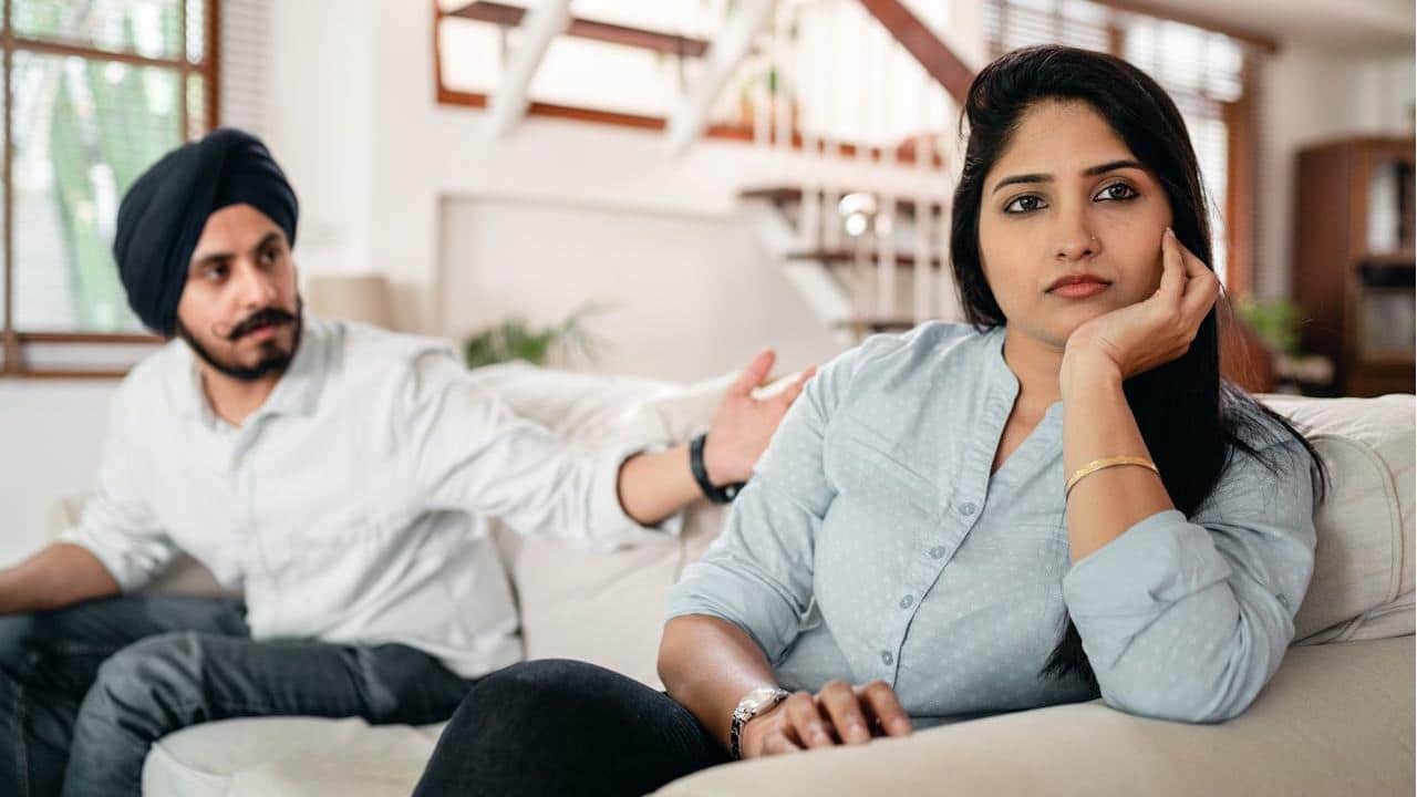 A woman is sitting on a couch with a distant expression while a man in a turban sits behind her, gesturing toward her.