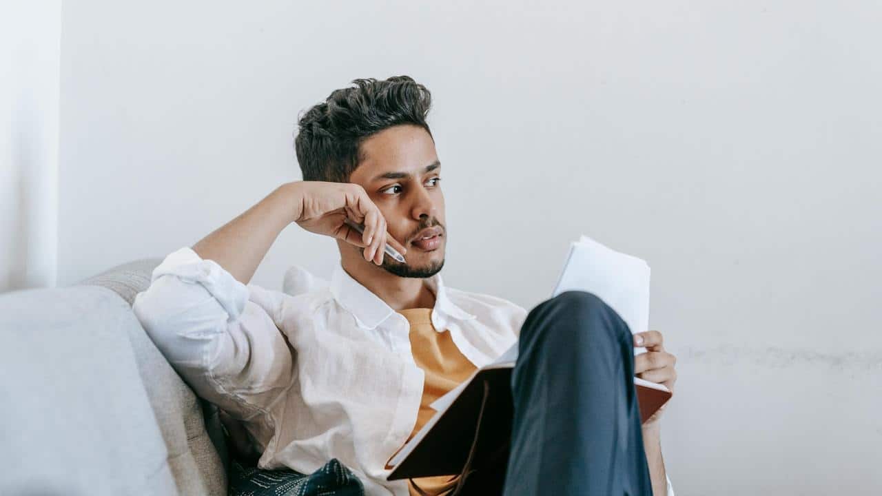 A man sitting on a couch, holding a notebook and looking thoughtfully to the side.