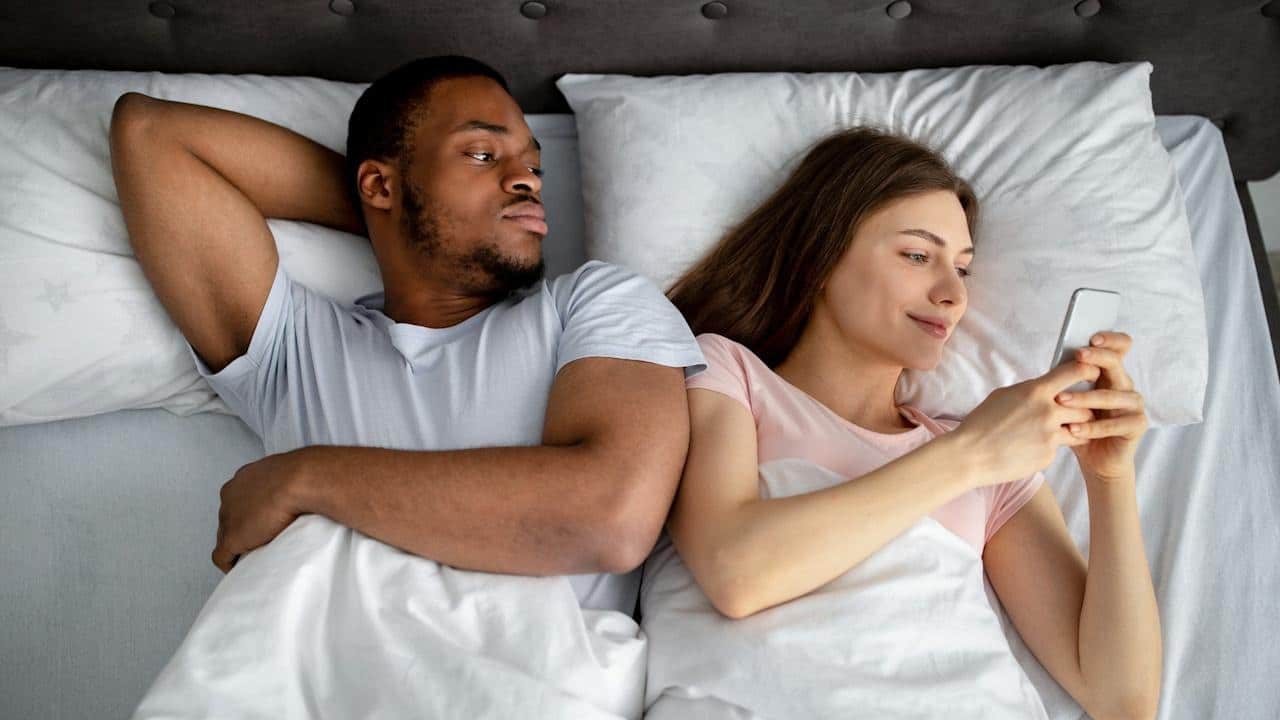 A man is lying in bed, looking suspiciously at the woman next to him who is smiling while looking at her phone.