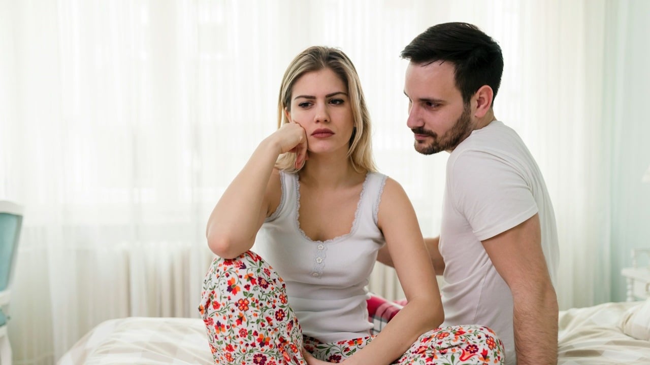 A sad woman is sitting on a bed, and her husband is looking at her while sitting next to her.
