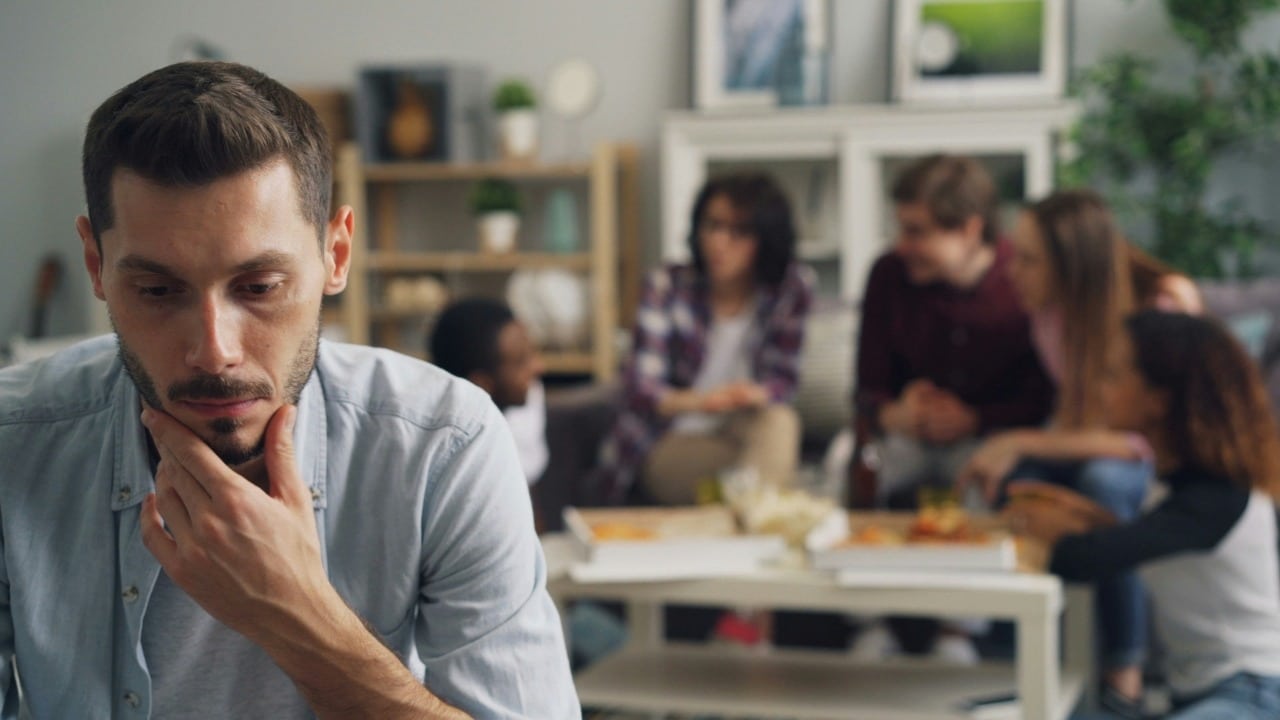 A man in the foreground looks down thoughtfully while a group of people talk behind him.
