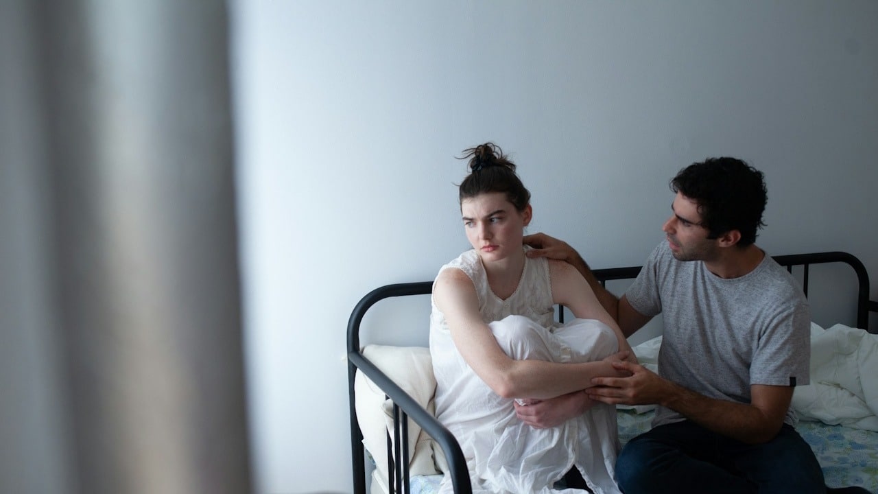 A man gently touches the shoulder of a sad or troubled woman sitting curled up on a metal bed frame.