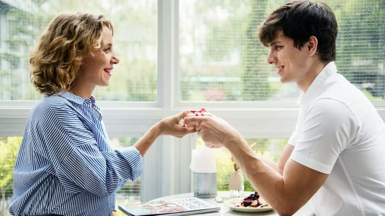 A smiling woman and man sit across a table, gently exchanging a small wrapped gift