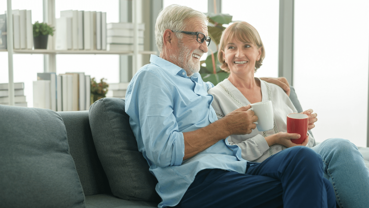 An elderly couple sits on a couch, smiling and holding mugs.