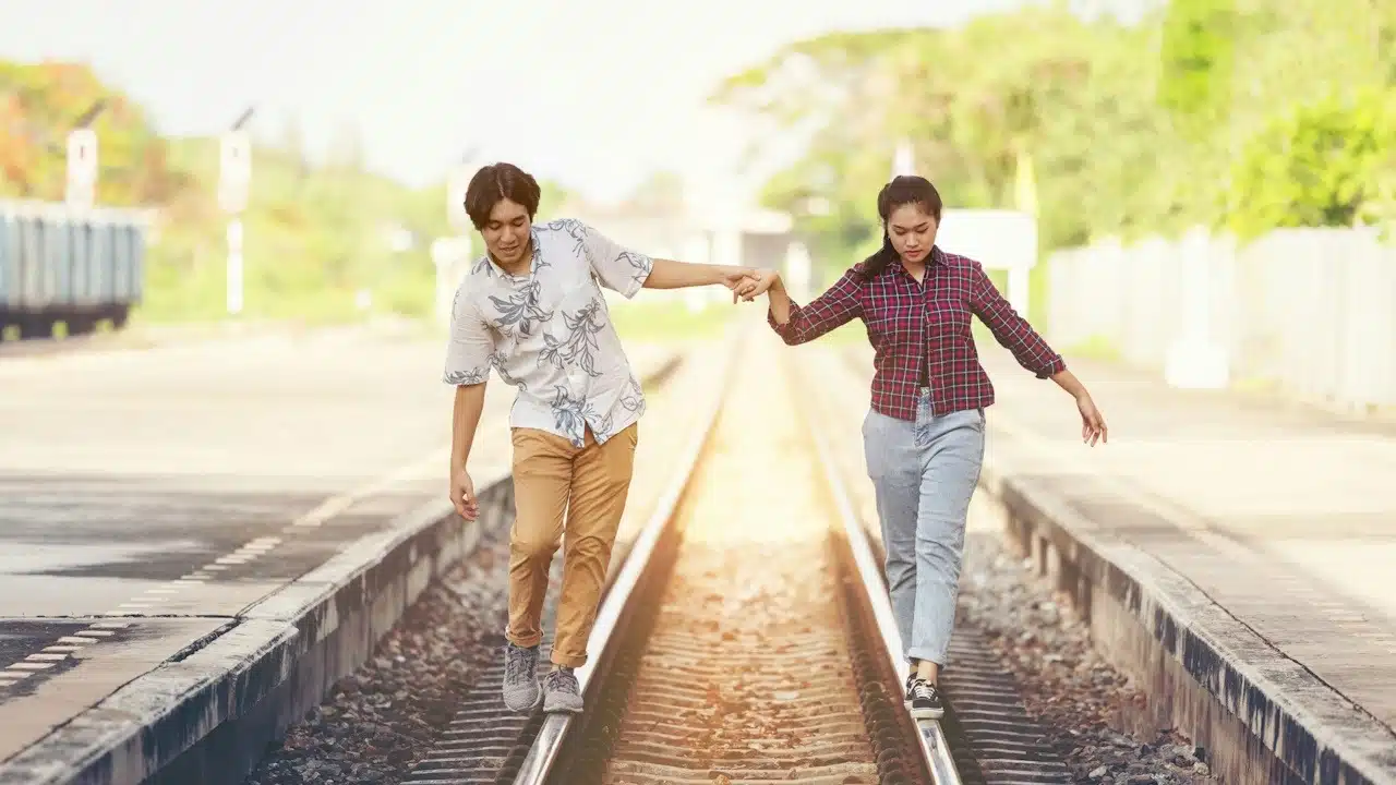 A couple walking on railway tracks while holding hands to maintain their balance at a train station.