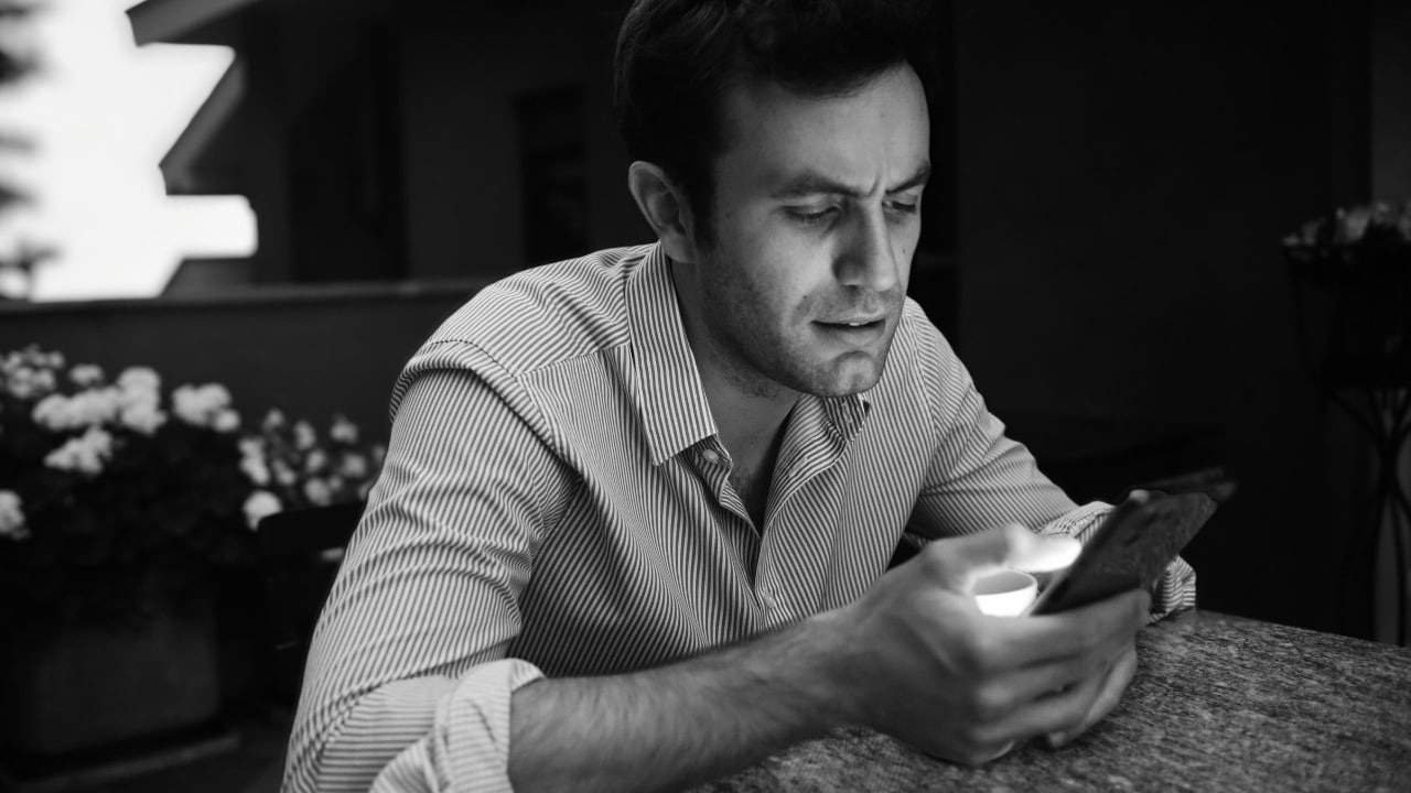 A black and white photo of a man looking down at a glowing smartphone screen.