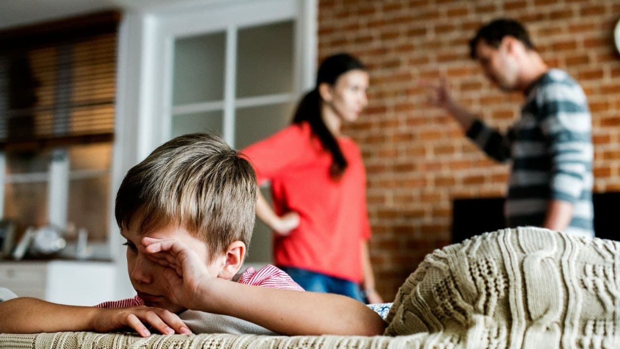 A young boy wipes his eyes while two adults stand blurred in the background.