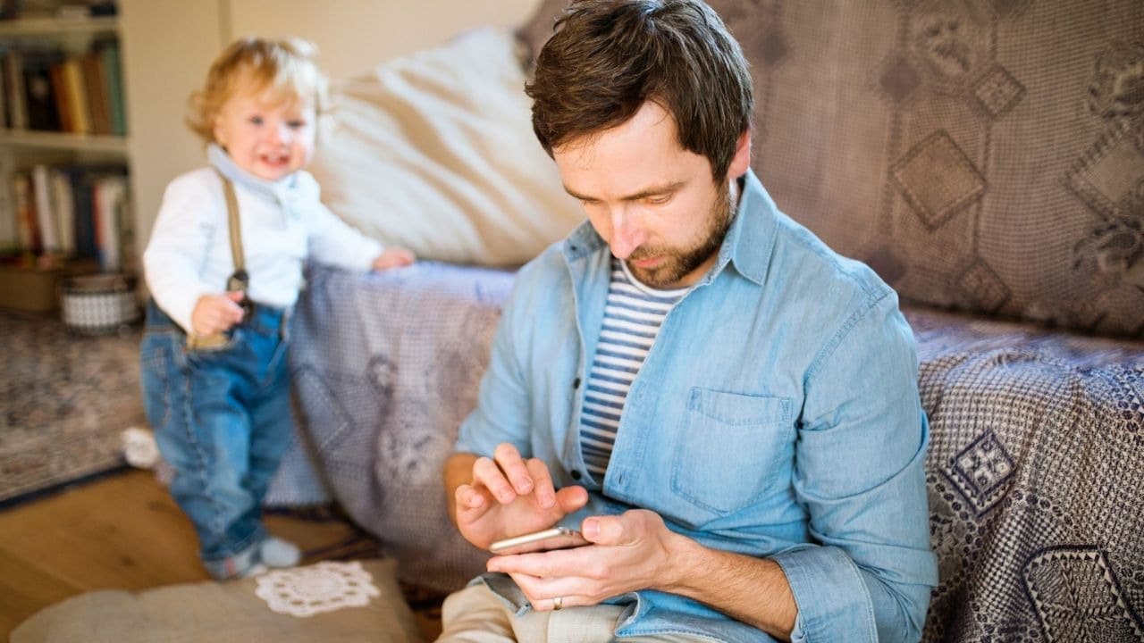A man is engrossed in his phone while a small child stands nearby in the background.