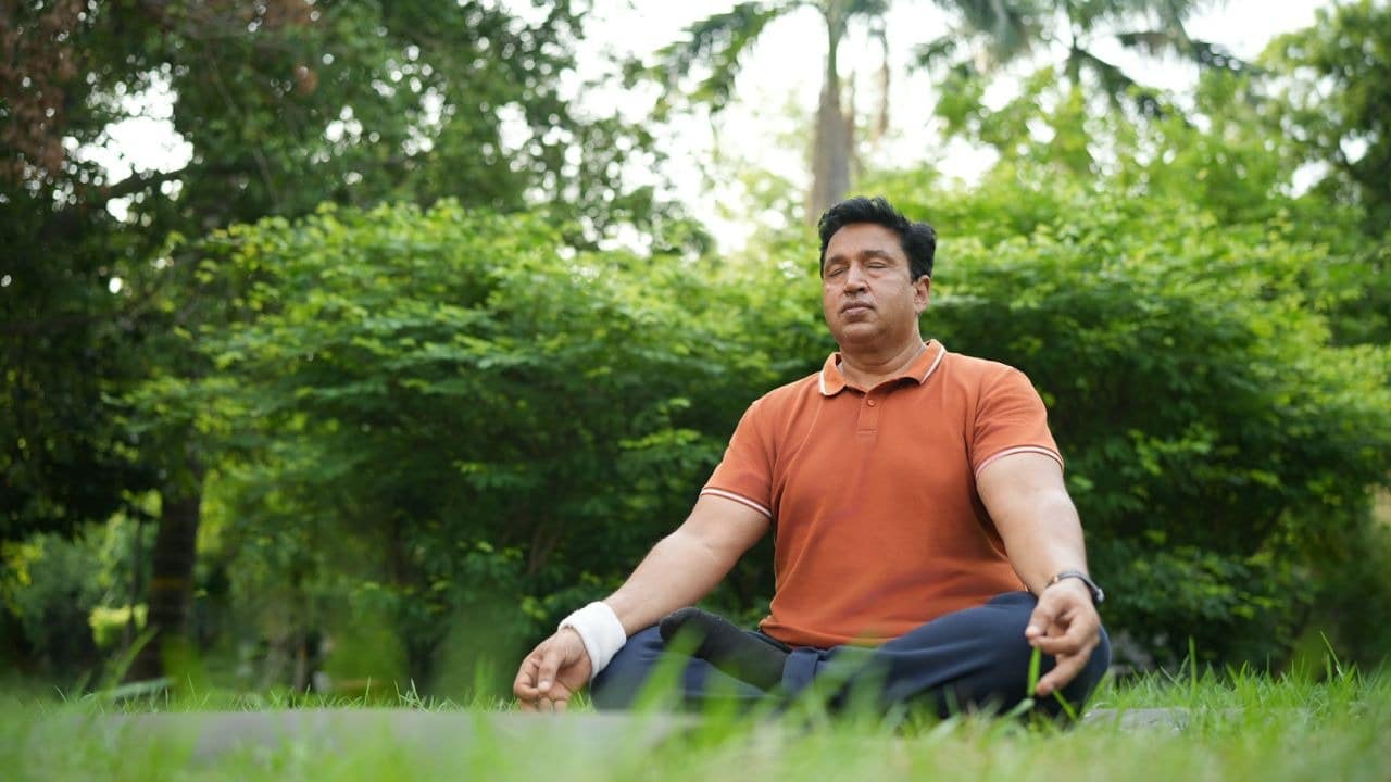 A serene man in an orange shirt sits cross-legged in the grass, meditating with his eyes closed.