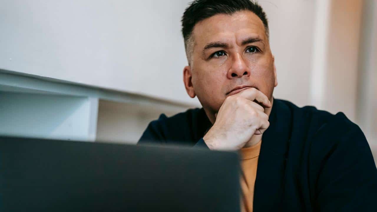 A man resting his chin on his hand, looking thoughtful while sitting at a desk.