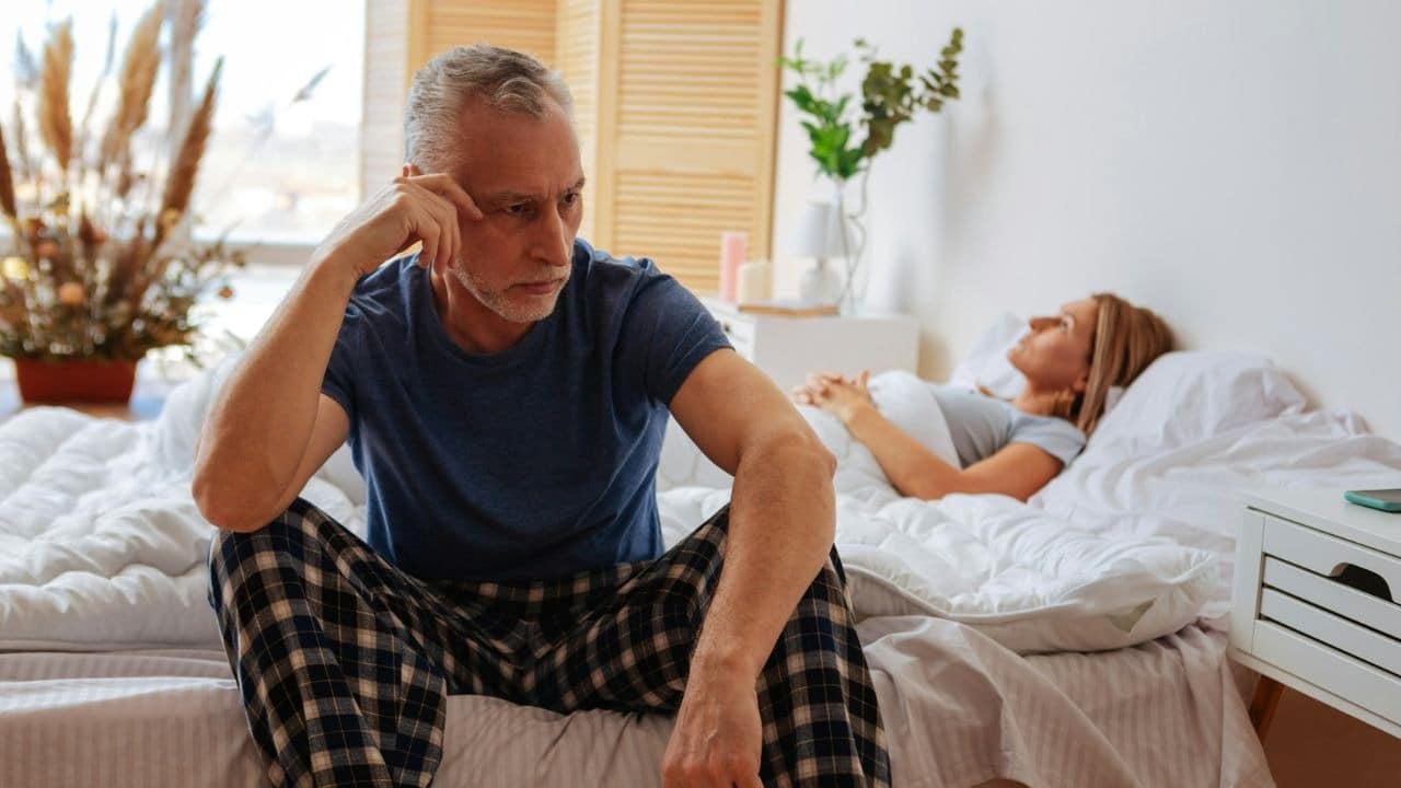 A mature man sits on a bed, looking troubled, with a woman sleeping behind him.