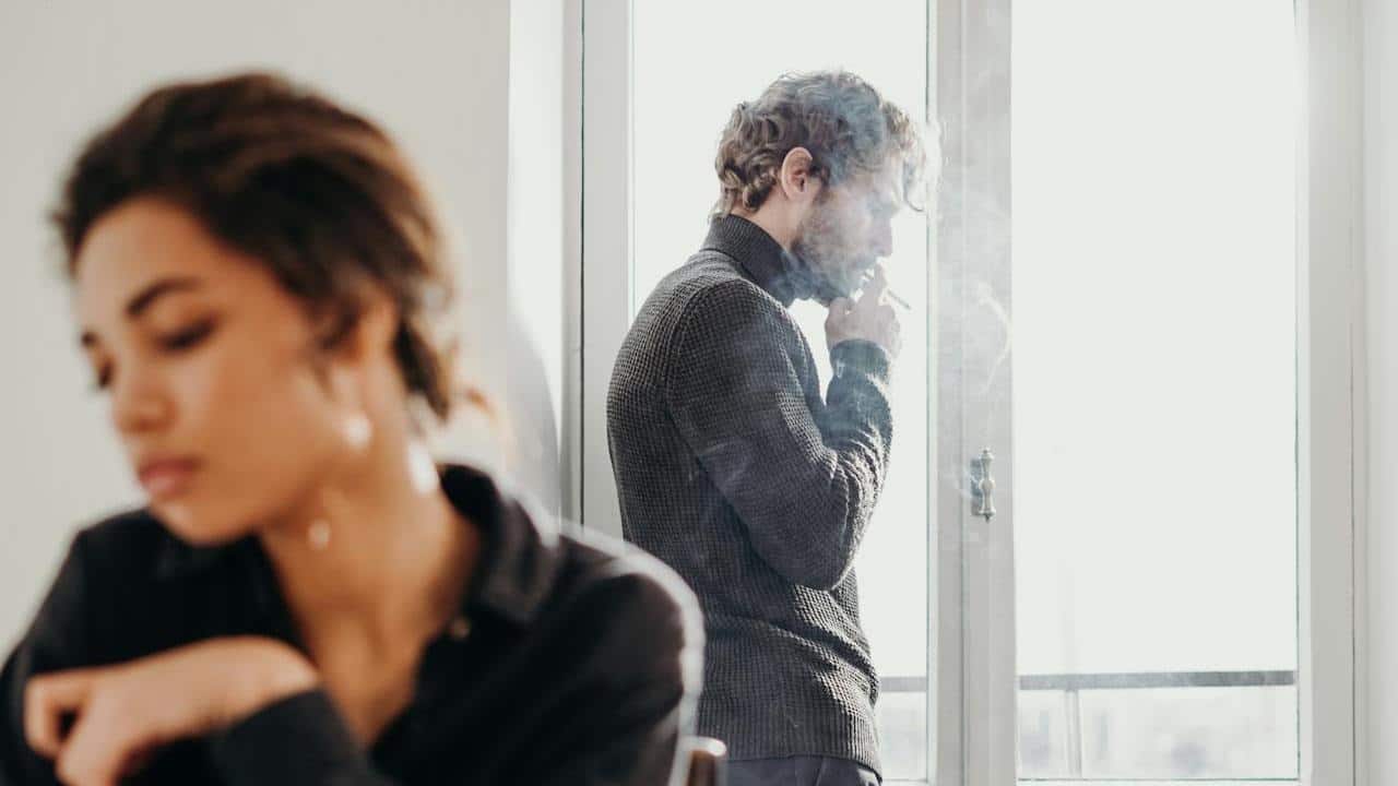 A man is standing in front of a bright window, smoking a cigarette, while a woman sits at a table in the blurred foreground.