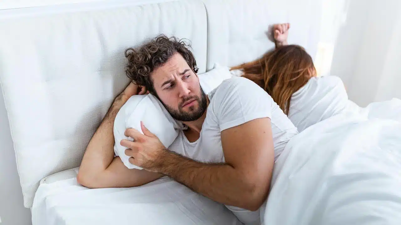 A man is lying in bed looking worried while a woman sleeps next to him with her back turned.