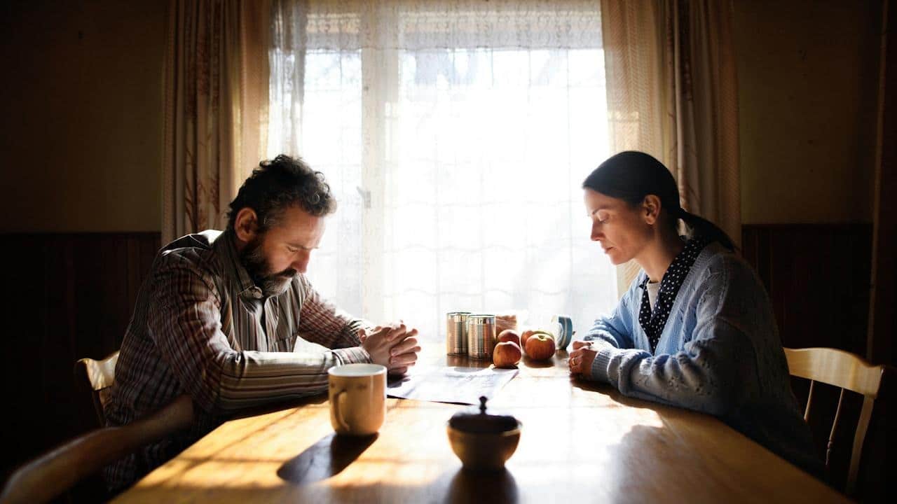A man and woman are sitting silently across from each other at a sunlit wooden table, both looking down.