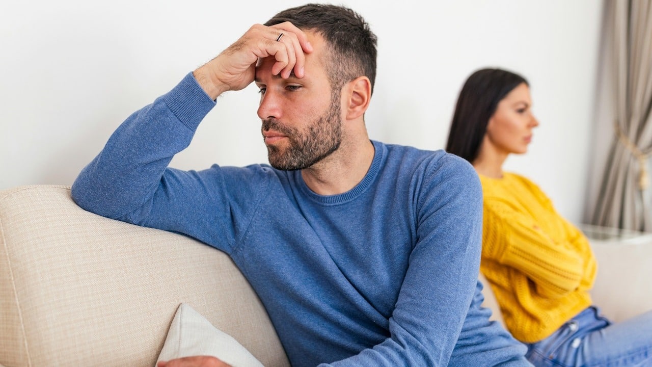 An upset man is holding his forehead with his hand as his wife sits behind him with crossed arms while they sit on a couch.