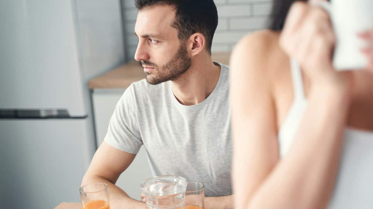 A man looks away in a kitchen, seemingly upset during a conversation with an out-of-focus woman.
