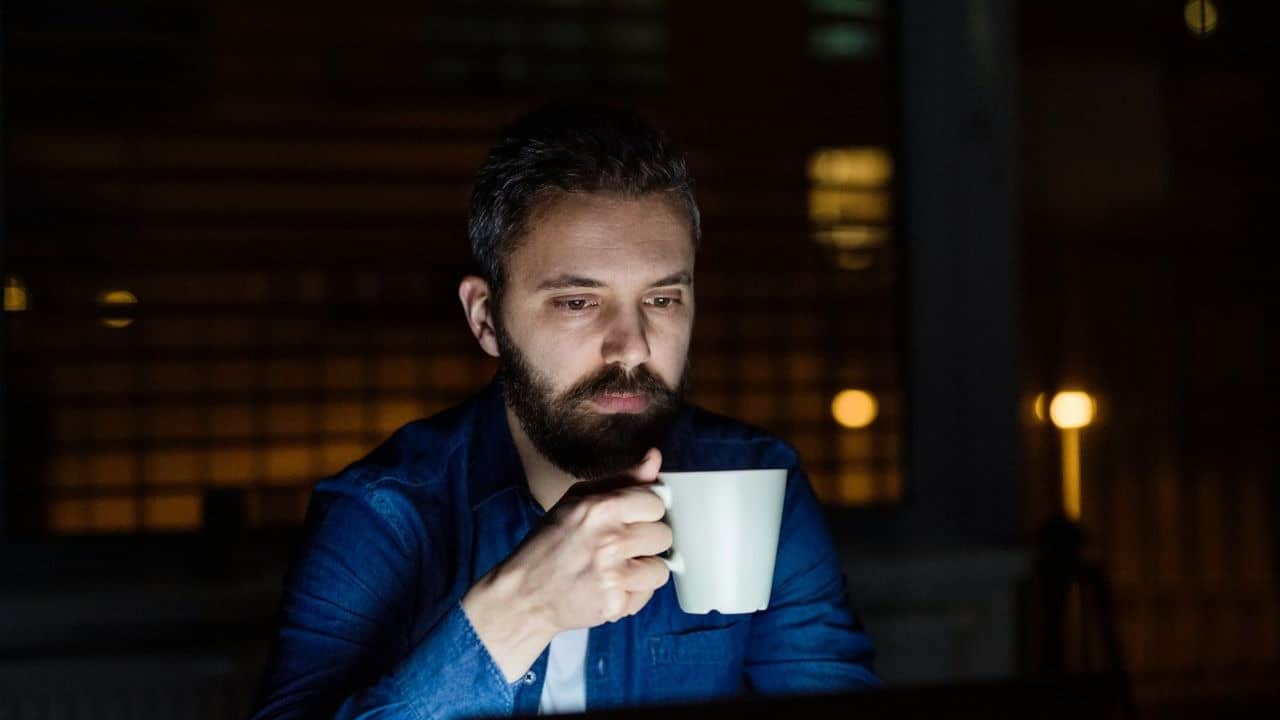 A man with a beard sits in the dark, holding a mug and looking at a screen.