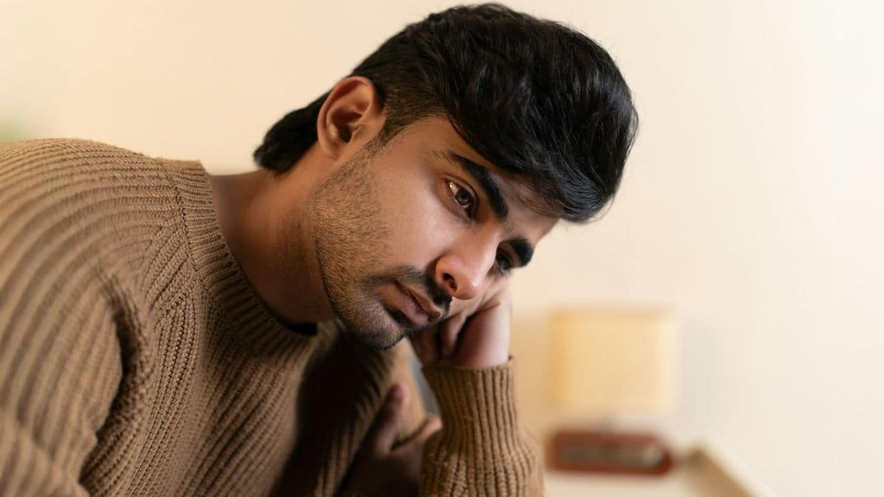 A man in a brown ribbed sweater rests his head on his hand and stares.