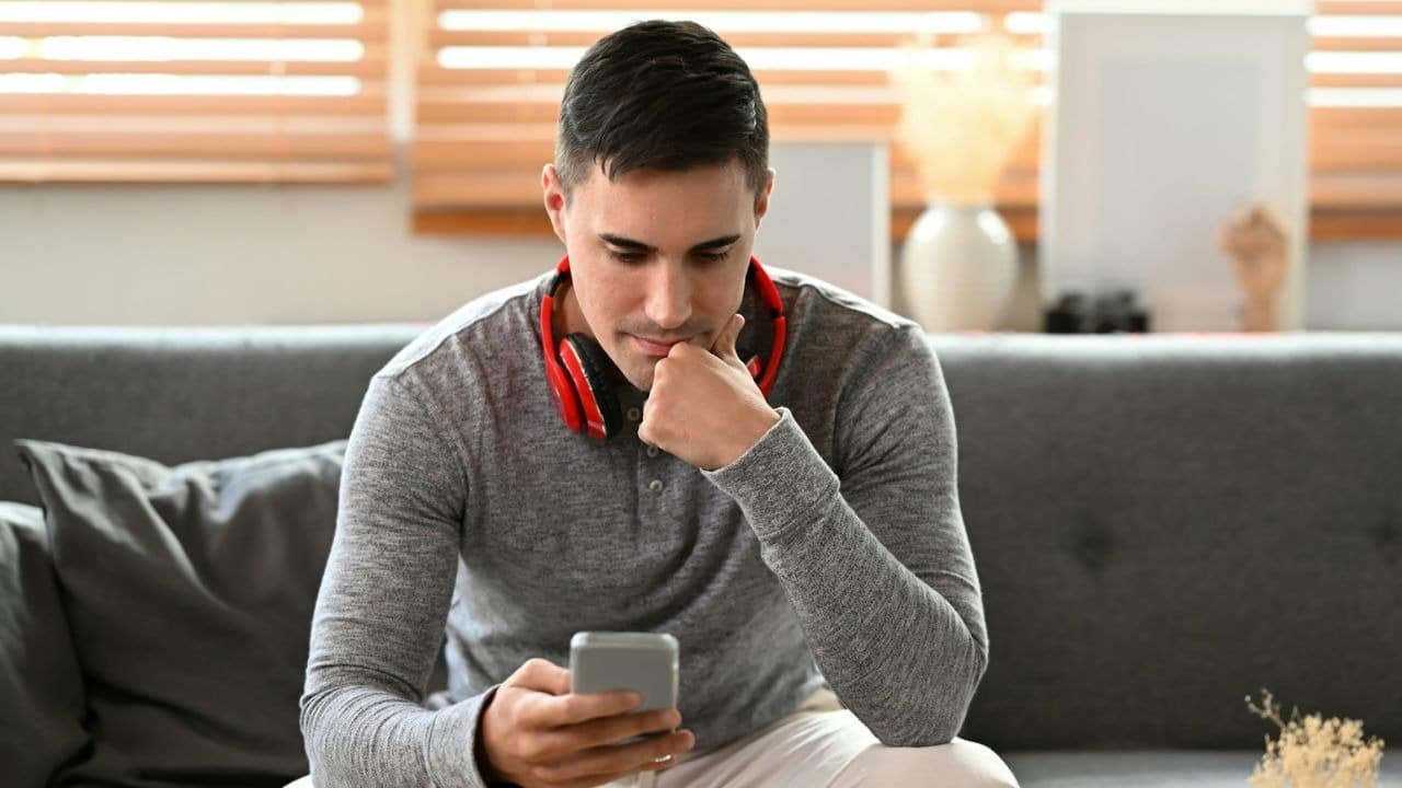 A man with red headphones around his neck looks down at a handheld smartphone screen.