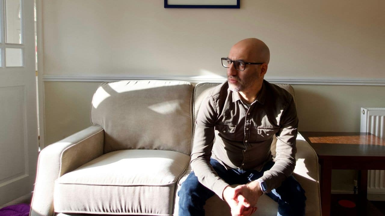 A bald man wearing glasses sits alone on a couch, looking to the side.