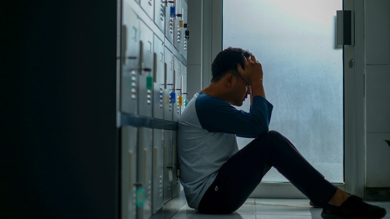 A man sitting on the floor while leaning on some lockers while holding his head in his hands.