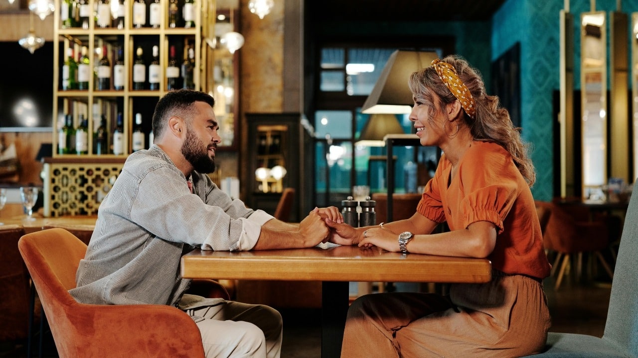 A man and woman sit at a cafe table holding hands and looking at each other.