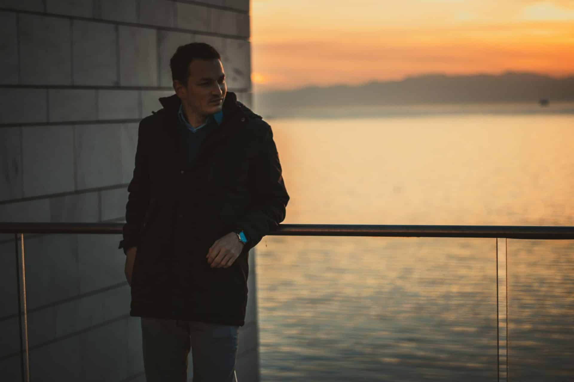 A man standing alone leaning against a railing at sunset.