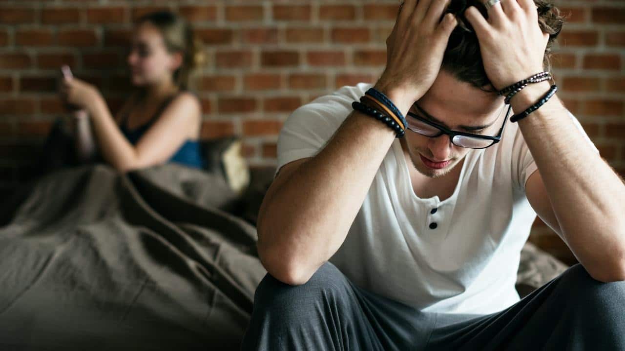 A woman is texting her ex while her worried husband is sitting holding his head with his hands.