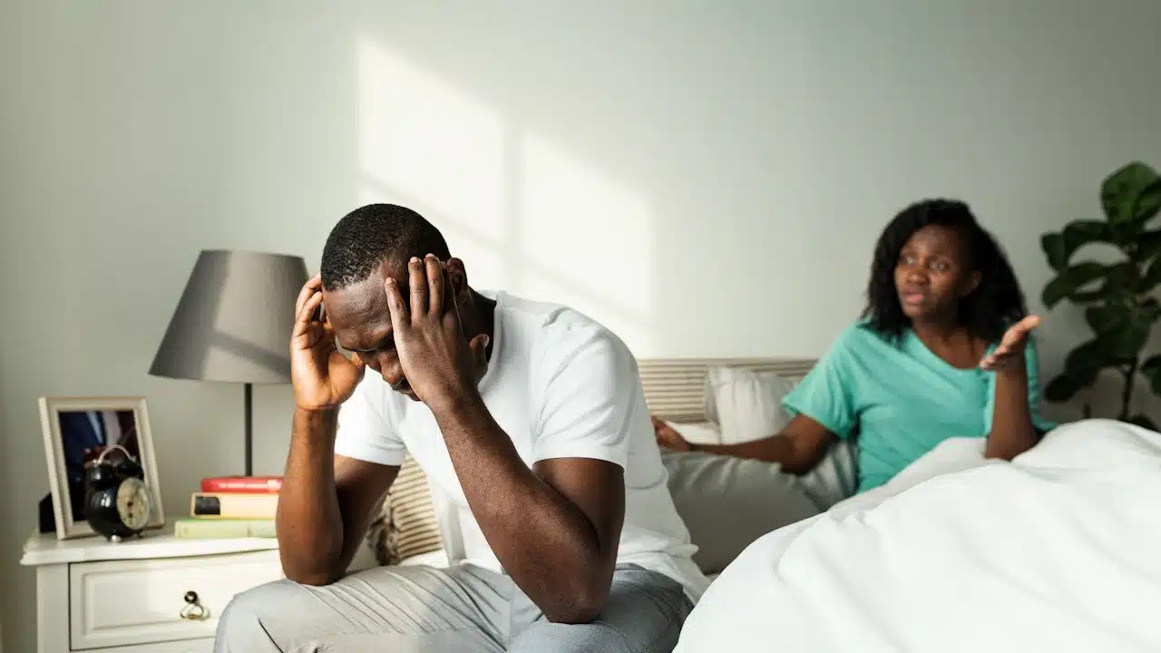 A woman arguing with a man who holds his head in his hands as they sit on a bed.