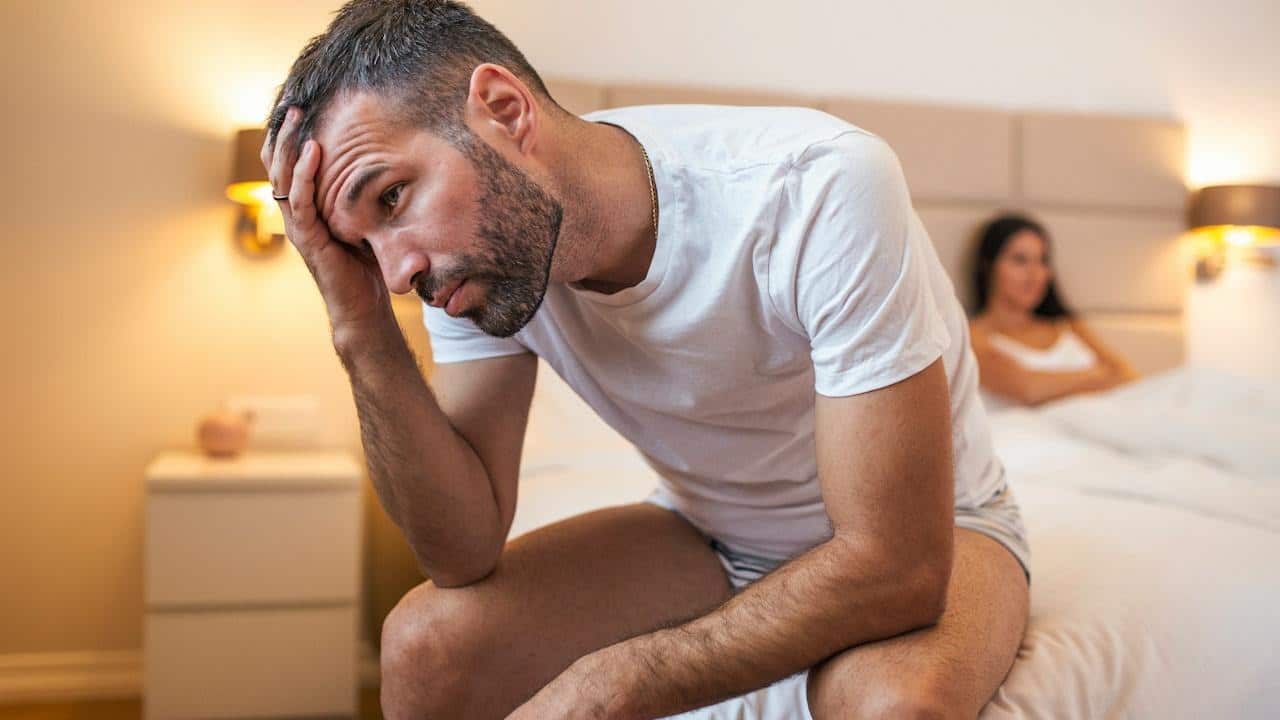 A man sitting on the edge of a bed looking distraught while a woman lies in the bed.