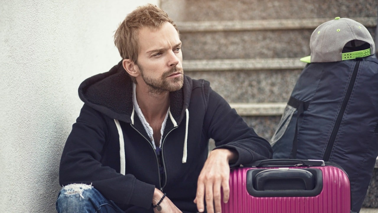A lightly bearded man resting his arm on a suitcase and sitting on some stairs while appearing lost in thought.