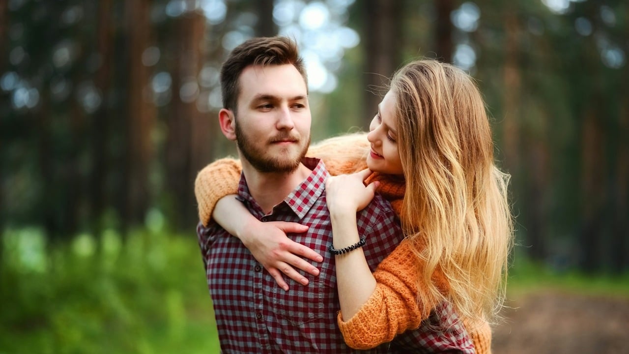 A bearded man carrying a woman on his back while they stand in some woods.