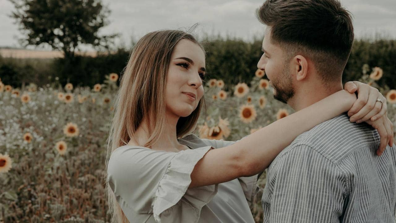 A woman staring in a man’s eyes while resting her arms on his arms as they stand in a sunflower field.
