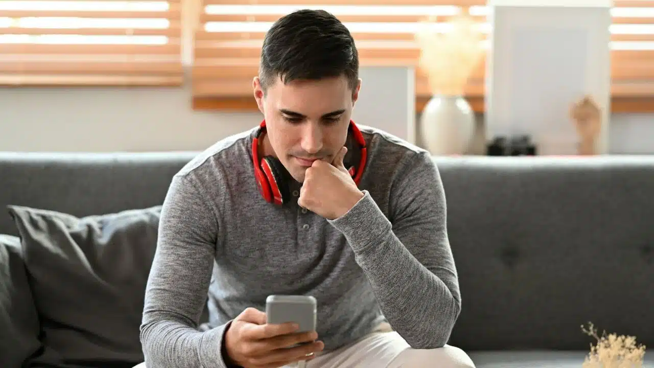 A man with red headphones around his neck looks down at a handheld smartphone screen.